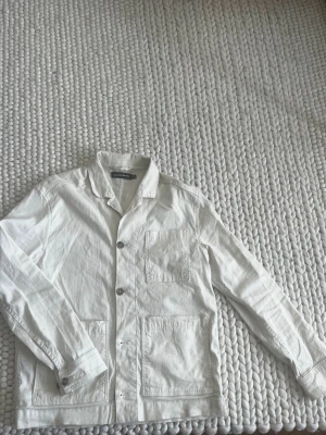 Vit overshirt från J.Lindeberg - Stilren vit overshirt från J.Lindeberg i linne. Klassisk krage, tre stora utanpåfickor och knäppning framtill med metallknappar. Perfekt lager på lager-plagg med avslappnad passform och raka ärmar.