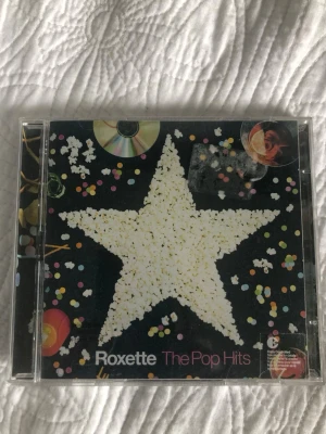 Roxette The Pop Hits CD - CD-album med Roxette, 'The Pop Hits', i plastfodral. Omslaget har en stor vit stjärna av popcorn mot svart bakgrund med färgglada konfettiprickar och CD-skivor. Baksidan listar 15 låtar, bland annat 'The Look', 'Joyride' och 'Stars'. Perfekt för dig som gillar popmusik.oöppnad 