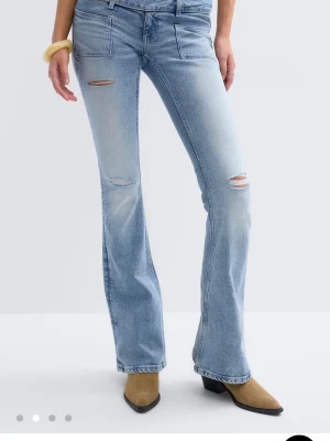 Ljusblå bootcut jeans med slitningar - Säljer ett par ljusblå bootcut jeans med slitna detaljer på låren och knäna. Jeansen har hög midja, stora framfickor och är tillverkade i mjuk denim. Perfekta för en avslappnad och trendig look.