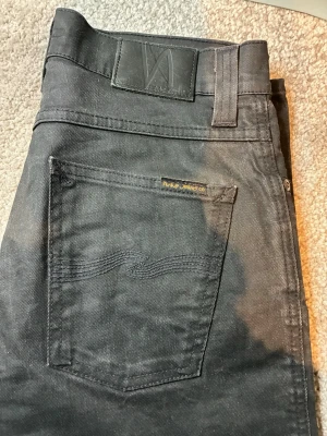Svarta jeans från Nudie Jeans - Snygga svarta jeans från Nudie Jeans med klassisk femficksmodell och diskret logga på bakfickan. Jeansen har raka ben och är tillverkade i stretchigt bomullsmaterial för extra komfort. Perfekta för en avslappnad och stilren look.