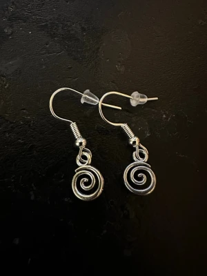 Silvriga spiralörhängen i metall - Unika örhängen i silverfärgad metall med spiralformad design. Hängande modell med krok och transparenta pluppar för extra säkerhet. Perfekta för dig som gillar detaljerade och kreativa accessoarer.