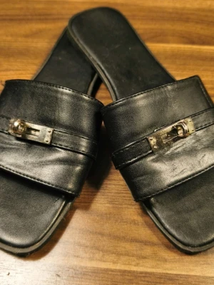 H Giulia Sandals  - Stylish black Giulia sandals in faux leather with a wide strap and unique metal buckle at the front. The sandals have a square toe and flat sole, which gives a modern vibe. Perfect for those who like minimalist design with a twist. Fits size 38-40