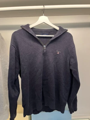 Mörkblå stickad tröja från Gant - Mörkblå stickad tröja från Gant med halv dragkedja och broderad logga på bröstet. Tröjan har ribbstickade muddar och krage, samt lång ärm. Perfekt för lager-på-lager och klassisk stil. 
