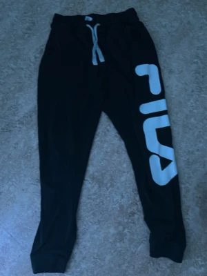 Svarta mjukisbyxor från Fila - Säljer ett par svarta mjukisbyxor från Fila med stort vitt och rött logotryck längs ena benet. Byxorna har resår i midjan med vit dragsko och muddar vid bensluten. Perfekta för chill eller träning.