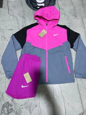 Nike träningsset rosa och grå - Snyggt träningsset från Nike med en färgstark vindjacka i rosa, grått och svart samt matchande rosa träningsshorts. Jackan har huva, dragkedja och två fickor med dragkedja. Shortsen har elastisk midja och vit Nike-logga. Perfekt för löpning eller gym.