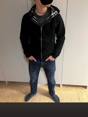 Burberry zip tröja -  Säljer en svart hoodie med dragkedja och huva. Insidan av huvan har ett rutigt foder i svart, vitt och rött. Hoodien har två fickor framtill och justerbara snören i huvan. Perfekt för en chill och avslappnad stil.