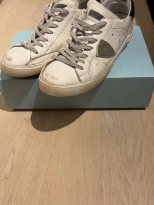 Vita Philippe Model sneakers - Snygga vita sneakers från Philippe Model med grå detaljer och gråa skosnören. Skorna har en låg silhuett och en ikonisk sköld på sidan. Ovandelen är i läder och hälen har en svart kontrastdetalj. Perfekta för en clean och trendig look.