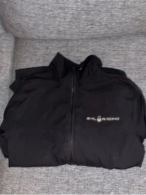 Svart vindjacka från Sail Racing - Snygg svart vindjacka från Sail Racing med dragkedja framtill och diskret logga på bröstet. Jackan har hög krage och är tillverkad i ett lätt, vindtätt material som passar perfekt för blåsiga dagar.