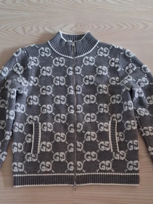 Grå ziptröja med GG-mönster Gucci stl L - Snygg grå ziptröja från Gucci med vitt GG-mönster över hela plagget. Tröjan har hög krage, dragkedja framtill och två fickor. Ribbstickade muddar vid ärmslut och nederkant samt detaljer i vitt. Perfekt för dig som gillar exklusiv streetwear. OBS! Sonen köpte denna tröja här på plick. Den är märkt stl S men den passade inte ens hans pappa som har stl M. Så väldigt stor i storleken, som en L. 