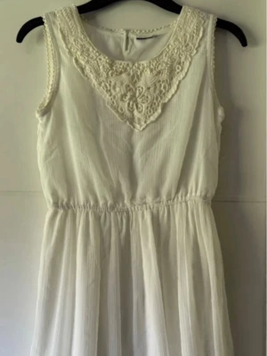Vit ärmlös klänning med spetsdetalj - Säljer en vit ärmlös klänning med foder och vacker spetsdetalj vid halsringningen och lätt rynkad midja. Klänningen har en luftig kjol och är perfekt för sommarens alla tillfällen. Materialet känns lätt och svalt. Kan passa till studenten. Bröst 88cm, längd 90cm.