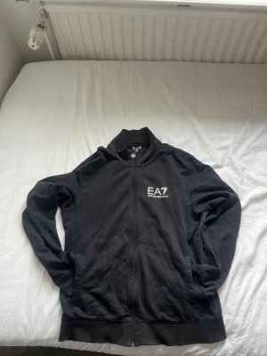 Svart zip hoodie från Emporio Armani - Snygg svart hoodie från Emporio Armani EA7 med dragkedja framtill och två fickor. Stort EA7-tryck på bröstet i vitt och rött. Klassisk passform och ribbade muddar. Perfekt för en stilren och sportig look.