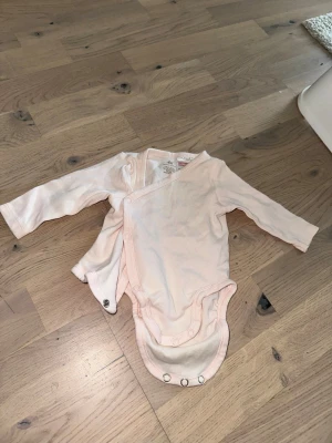 Ljusrosa omlottbody från H&M - Söt ljusrosa omlottbody med långa ärmar från H&M. Tillverkad i mjuk ekologisk bomull och har tryckknappar längs sidan och i grenen för enkel påklädning. Perfekt för små barn och bebisar.