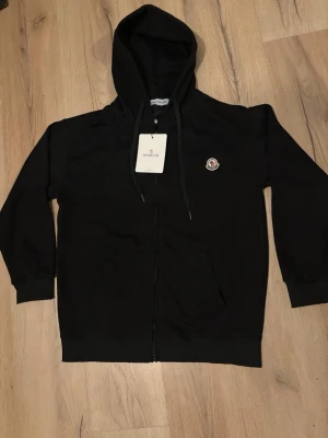 Moncler zip hoodie - Svart hoodie från Moncler med dragkedja framtill, huva med dragsko och klassisk Moncler-logga på bröstet. Tillverkad i mjukt material som känns skönt mot huden. Perfekt för en avslappnad och stilren look. Storlek XL men passar även L