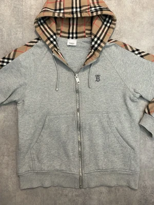 Grå Burberry hoodie  - Snygg grå hoodie från Burberry med dragkedja, klassiskt beige-rutigt mönster på huva och ärmar samt broderad B-logga på bröstet. Tillverkad i mjuk bomullsblandning och har kängurufickor framtill. Perfekt för en trendig och avslappnad stil.
