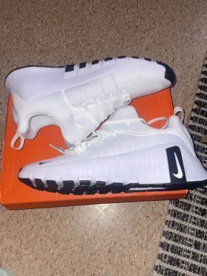 Vita Nike sneakers med svarta detaljer - Snygga vita Nike sneakers med svarta detaljer och logga på sidan. Skorna har en låg profil, mesh- och syntetmaterial samt robust sula med svart mönster undertill. Perfekta för dig som gillar sportig och clean stil.