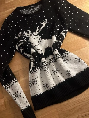 Svartvit stickad tröja med renmotiv - Stickad tröja i svart och vitt med stort renmotiv framtill, snöflingor och prickar som ger vinterkänsla. Rund halsringning och långa ärmar. Perfekt för dig som gillar mysiga vinterplagg med grafiskt mönster.