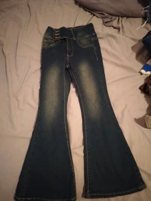 Mörkblå bootcut jeans med hög midja - Säljer ett par mörkblå bootcut jeans med hög midja och dubbla knappar i linningen. Jeansen har klassiska fickor fram och bak samt snygga slitningar längs benen för en trendig look. Perfekta för dig som gillar retrostil och vill ha ett par jeans som sticker ut.