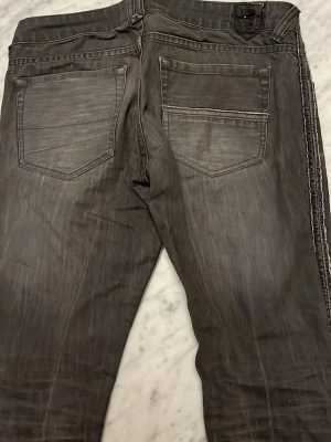 Grå slitna jeans med revär - Säljer ett par grå jeans med slitna detaljer och revär längs sidorna. Jeansen har fem fickor och en klassisk knappgylf. Materialet är troligtvis bomull med stretch för extra komfort. Perfekta för en avslappnad och trendig look.