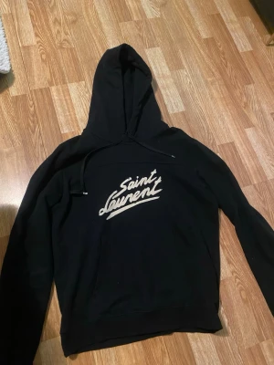 Svart Saint Laurent hoodie - Svart hoodie från Saint Laurent med vit logotyp på bröstet. Klassisk huva med dragsko och långa ärmar. Perfekt för en avslappnad och trendig stil. Tillverkad i mjukt material för extra komfort. Storlek M jag är 182 väger 78 skriv för mer info 