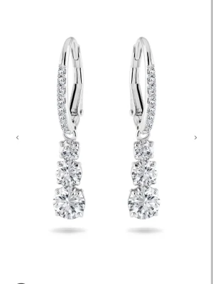 Silverörhängen med gnistrande stenar Swarovski  - Säljer ett par eleganta örhängen i silverfärg med tre runda, glittrande stenar i rad och små stenar längs bågen. Perfekt för dig som gillar stilrena och lyxiga accessoarer. Kommer i en söt rosa ask.