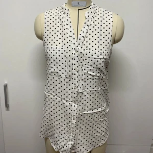 Vit ärmlös blus med svarta prickar - Säljer en vit ärmlös blus med svarta prickar och knäppning framtill. Blusen har en avslappnad passform och V-ringning utan krage. Perfekt för en trendig och luftig stil under varmare dagar. Som ny.