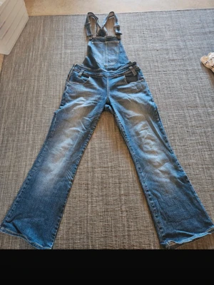 Blå denim hängselbyxa med utsvängda ben - Säljer en snygg blå denim jumpsuit/hängselbyxa med klassiska hängslen och utsvängda ben. Byxan har flera fickor fram och bak, justerbara axelband. De är väldigt stretchiga och bekväma i materialet 