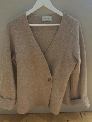 Beige kofta från Neo Noir - Mysig beige kofta från Neo Noir med djup V-ringning och en knapp framtill. Koftan har långa ärmar och är tillverkad i ett mjukt, stickat material. Perfekt för lager på lager och enkel att matcha med olika outfits. pris kan diskuteras 