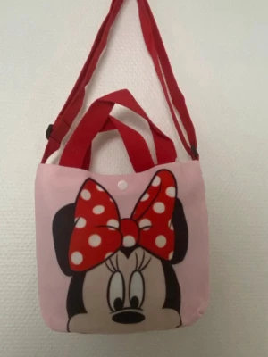 Söt Disney barn handväska / axelväska   - Handväska med två olika Disney-motiv: Minnie Mouse med röd prickig rosett på ena sidan och Nalle Puh med Nasse på andra. Väskan är i tyg, har röda handtag och axelrem. Perfekt för dig som gillar gulliga accessoarer och vill ha något unikt. 125kr/st , helt ny 