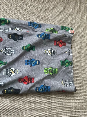 Halsduk  - Grå t-shirt från halsduk  med färgglatt Lego Ninjago-tryck över hela plagget. Motiven är i blått, grönt, rött, svart och vitt. Tillverkad i mjuk bomull som är skön att ha på sig. Perfekt för dig som gillar Lego och vill ha en unik och lekfull stil.