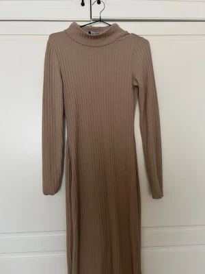 Beige ribbad långklänning från Pieces - Långklänning i beige från Pieces med ribbad struktur och lång ärm. Klänningen har en hög krage och är figurnära i modellen, perfekt för en stilren och enkel look. Tillverkad i mjukt material som känns skönt mot huden.💕