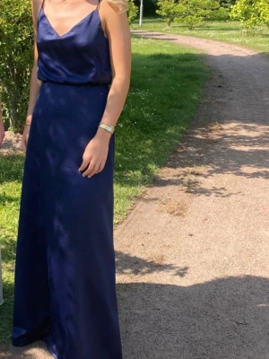 Marinblå balklänning  - Säljer en elegant marinblå långklänning i glansigt satinmaterial. Klänningen har smala axelband, v-ringad urringning och en lös, följsam passform som ger en stilren siluett. Perfekt för speciella tillfällen!