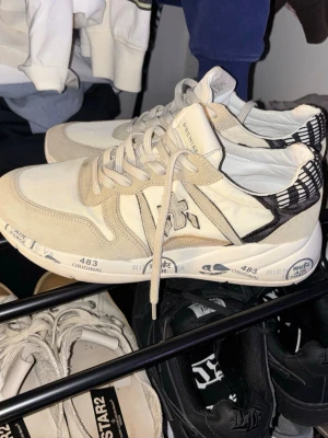 Premiata vita - Snygga vita sneakers från Premiata med beige detaljer och svartvitt mönster på hälen. Skorna har en chunky sula med tryck och klassisk snörning. Tillverkade i en mix av mocka och textil.