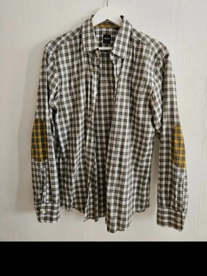 Rutig skjorta från BOSS med lappar - Rutig skjorta från BOSS i bomull med klassisk krage och knappar framtill. Skjortan har lång ärm och kontrasterande lappar i gult på armbågarna för en cool detalj. Perfekt för dig som gillar en avslappnad men ändå stilren look.