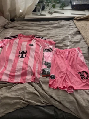 Rosa fotbollskit Inter Miami CF - Säljer ett rosa Messi fotbollssätt från Inter Miami CF med matchtröja och shorts. Tröjan har svarta ränder på axlarna, klubbmärke och tryck på bröstet. Shortsen är helrosa med nummer 10 och klubbmärke. Perfekt för dig som älskar fotboll och vill sticka ut på planen.strjmporna är rosa i storlek M.