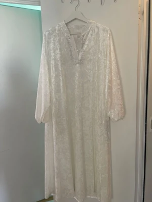 Vitt långt nattlinne med knappar - Långt vitt nattlinne i mjukt, glansigt tyg med diskret mönster. Har långa ärmar, lös passform och knappar vid halsringningen. Perfekt för mysiga kvällar hemma. Passar mellan storlek S och M