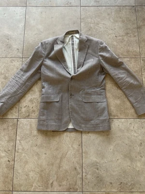 Ljusgrå kavaj - Snygg ljusgrå kavaj med två knappar framtill och klassiska slag. Kavajen har två fickor med lock och en innerficka. Tunt randigt foder på insidan. Perfekt för en stilren look och passar till många olika outfits.