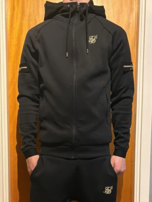 Svart hoodie set med byxor  från SikSilk med dragkedja - Svart hoodie från SikSilk med hel dragkedja framtill, justerbar huva med snören och guldfärgad logga på bröstet. Ärmarna har diskreta ränder och muddar vid handlederna. Perfekt för en sportig och avslappnad stil. 
