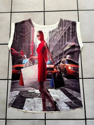 Topp med New York-print och shoppingmotiv storlek L/XL - Cool ärmlös topp med färgstarkt tryck av en kvinna i röd klänning och shoppingpåsar på en New York-gata. Basen är vit och materialet känns som mjuk polyester. Perfekt för dig som gillar statement-plagg och unika motiv.