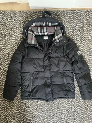 Svart pufferjacka från Burberry - Snygg svart pufferjacka från Burberry med avtagbar huva och klassiskt rutigt foder i grått, vitt och rött. Jackan har två dragkedjefickor fram och en patch med 'BURBERRY LONDON ENGLAND' på ärmen. Perfekt för kalla vinterdagar. 