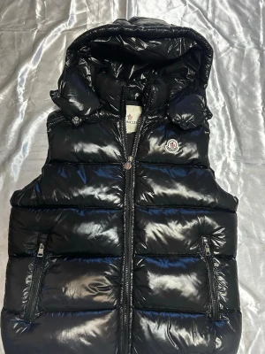 Svart dunväst från Moncler - Snygg svart dunväst från Moncler med glansigt material och avtagbar huva. Västen har två dragkedjeförsedda fickor och Moncler-logga på bröstet. Perfekt för lager-på-lager och ger en cool look till din outfit.
