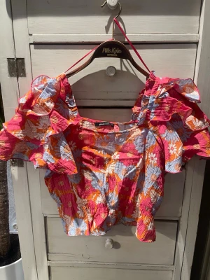 Blommig croppad topp med volang - Färgstark croppad topp från Gina Tricot med blommigt mönster i rosa, orange och ljusblått. Toppen har stora volangärmar och fyrkantig ringning. Materialet är lätt och krinklat, perfekt för sommarens alla tillfällen.