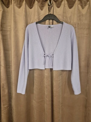 Ljuslila ribbad kofta från H&M - Trendig ljuslila ribbad kofta från H&M med croppad passform och långa ärmar. Koftan har en djup V-ringning och knytdetalj framtill som ger en snygg och avslappnad look. Perfekt att slänga över ett linne eller topp för en chill vibe.