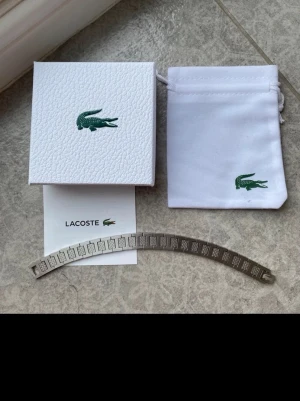 Silverfärgat armband från Lacoste - Snyggt oanvänt armband i silverfärgad metall från Lacoste den är 20cm lång men kan enkelt justeras, Kommer med original ask och tygpåse.