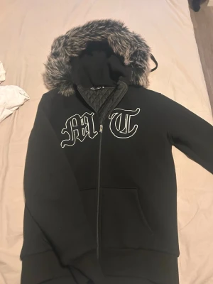 Svart hoodie med fuskpäls och tryck - Svart hoodie från Active Boys med dragkedja, stor huva med fuskpäls och quiltat innerfoder. Framsidan har vita bokstavstryck 'MT' i gotisk stil. Perfekt för dig som vill ha en streetwear-look med extra värme. Sitter hur bra som helst i pass form även om du har lite längre armar