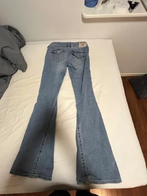 Ljusblå bootcut jeans från True Religion - Säljer ett par ljusblå bootcut jeans från True Religion med klassiska fickor och broderade detaljer bak. Jeansen har låg midja och utsvängda ben, perfekt för en retroinspirerad look. Skriv innan du köper bara så jag vet❤️
