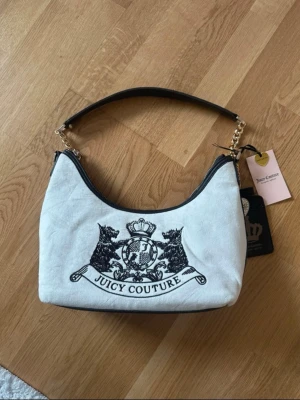 Vit Juicy Couture handväska i sammet - Snygg vit handväska från Juicy Couture i mjuk sammet med svart broderad logga och emblem framtill. Väskan har guldfärgade kedjedetaljer på axelremmen och svarta detaljer. Perfekt accessoar för att lyfta din outfit.