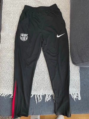 Svarta Nike Barcelona träningsbyxor - Snygga svarta träningsbyxor från Nike med FC Barcelona-logga på vänster ben och vit Nike Swoosh på höger ben. Byxorna har röda detaljer vid bensluten och är tillverkade i ett lätt och stretchigt material, perfekta för fotbollsträning eller chill.