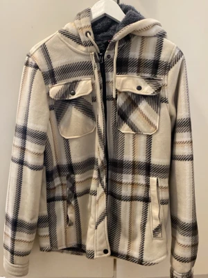 Beige och brun rutig overshirt med huva - Snygg overshirt i beige, brunt och svart rutigt mönster. Jackan har huva, två bröstfickor med lock och knappar samt dragkedja framtill. Mjuk och varm fleece-liknande material, perfekt för lager på lager. Passar dig som gillar avslappnad och trendig stil.
