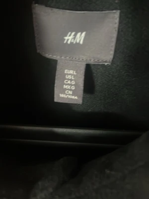 Svart overshirt från H&M - Stilren svart overshirt från H&M med klassisk krage och knäppning framtill. Två stora bröstfickor ger en cool och avslappnad look. Perfekt att bära över en t-shirt för en trendig lager-på-lager-stil.