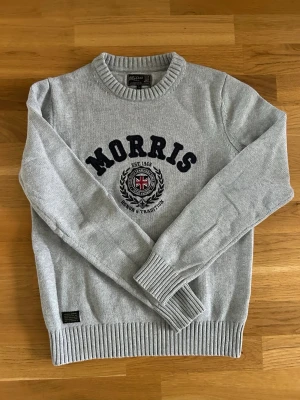 Grå stickad tröja från Morris - Snygg grå stickad tröja från Morris med rund halsringning och ribbade muddar. Framtill står det 'Morris' i svart text. Jättebra skick!